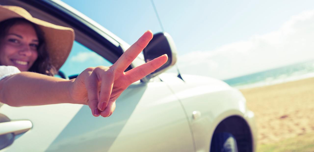 A girl in a brimmed hat, riding in a car, flashing a peace sign with her fingers while parked on a beach